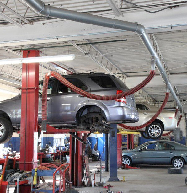 Vehicles shown lifted in bays in an auto repair shop while overhead vehicle exhaust extraction systems are installed to removal contaminants.